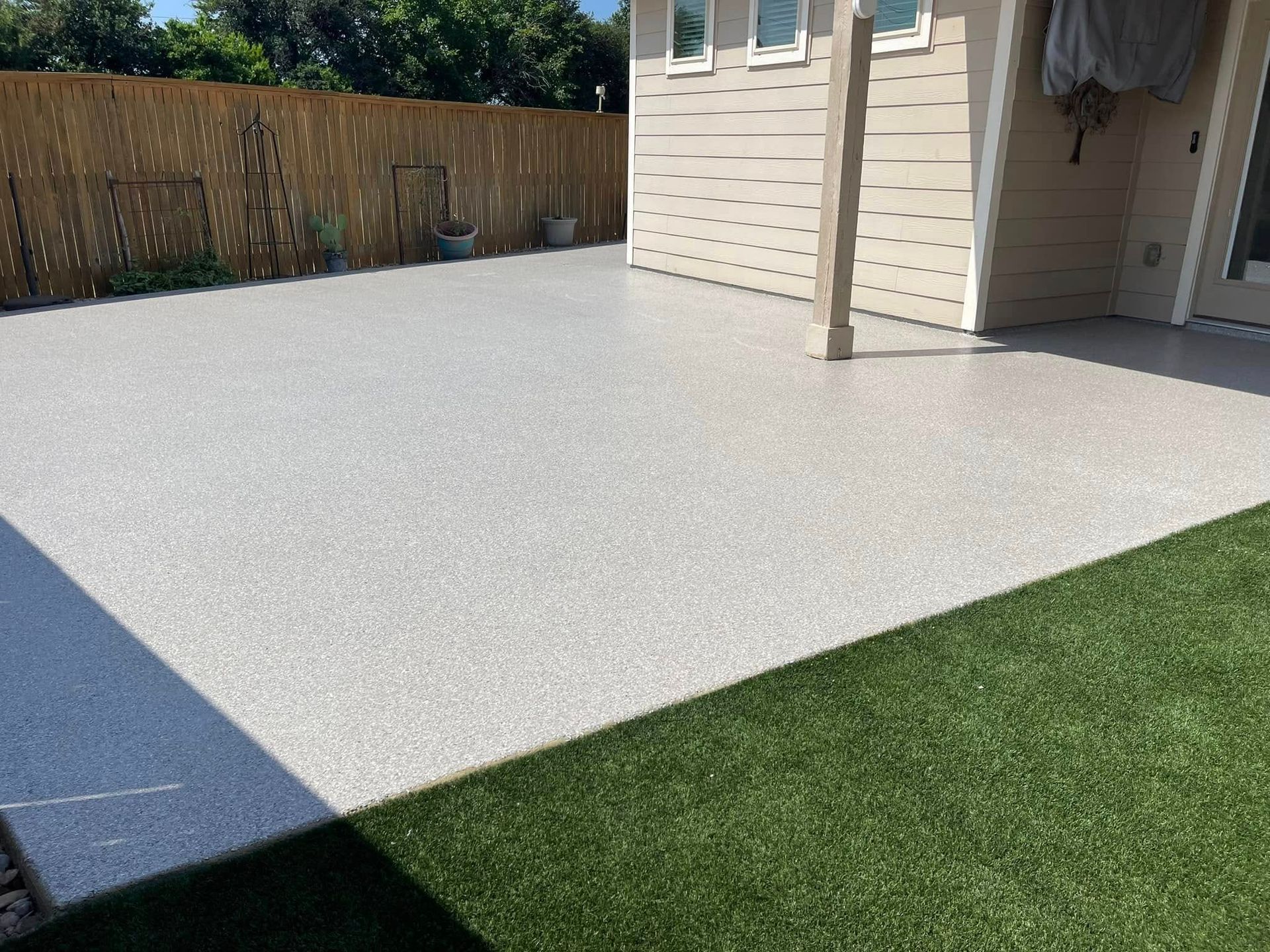 Concrete patio with speckled surface, next to green artificial grass. House and wooden fence in background.