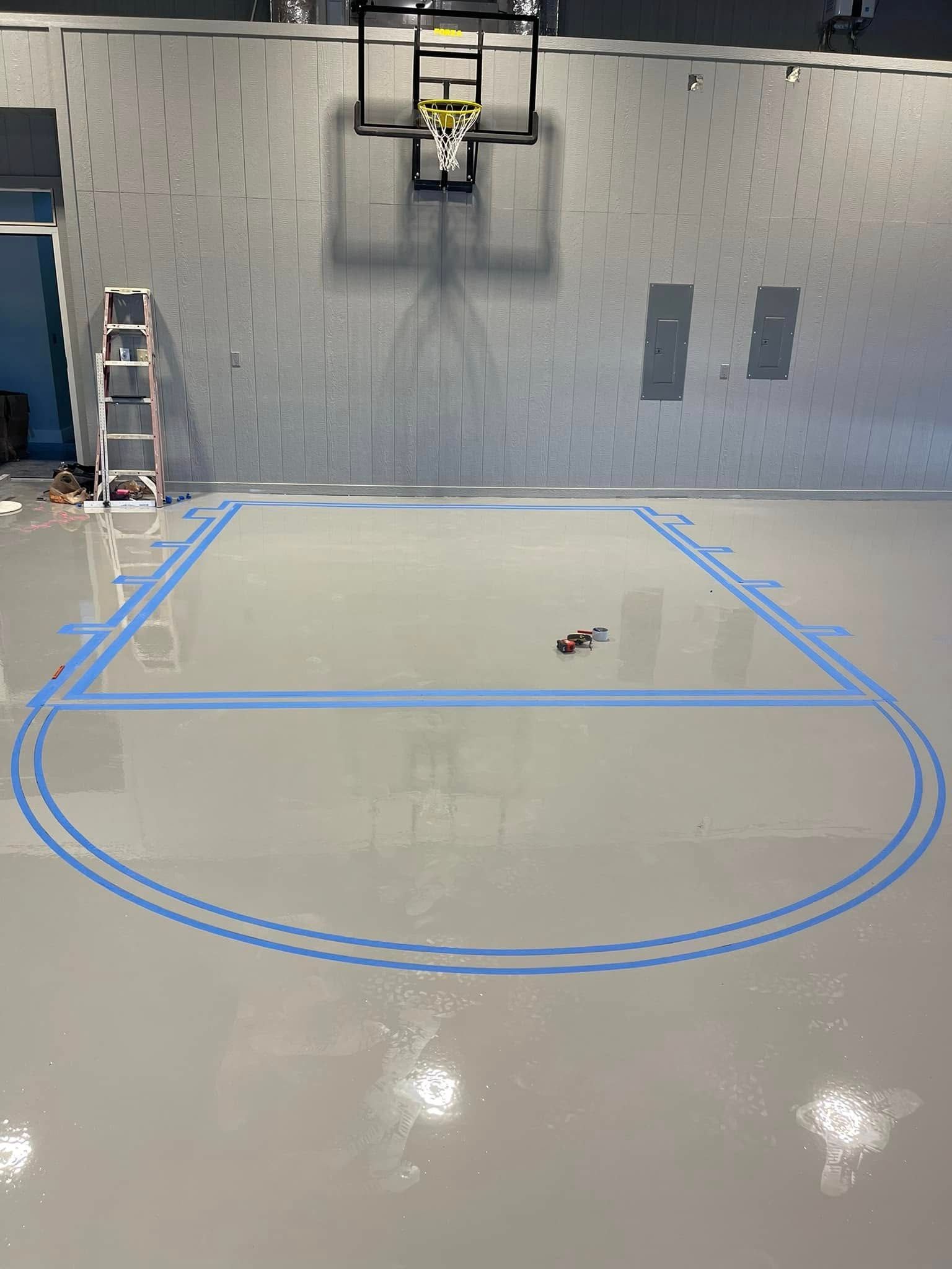 A basketball court floor being taped with blue tape, under a hoop, in a gray-walled gym.