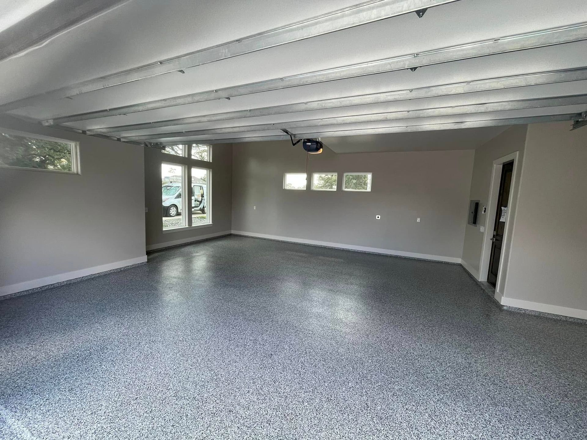 A spacious, empty garage with a speckled epoxy floor, white ceiling beams, and windows.