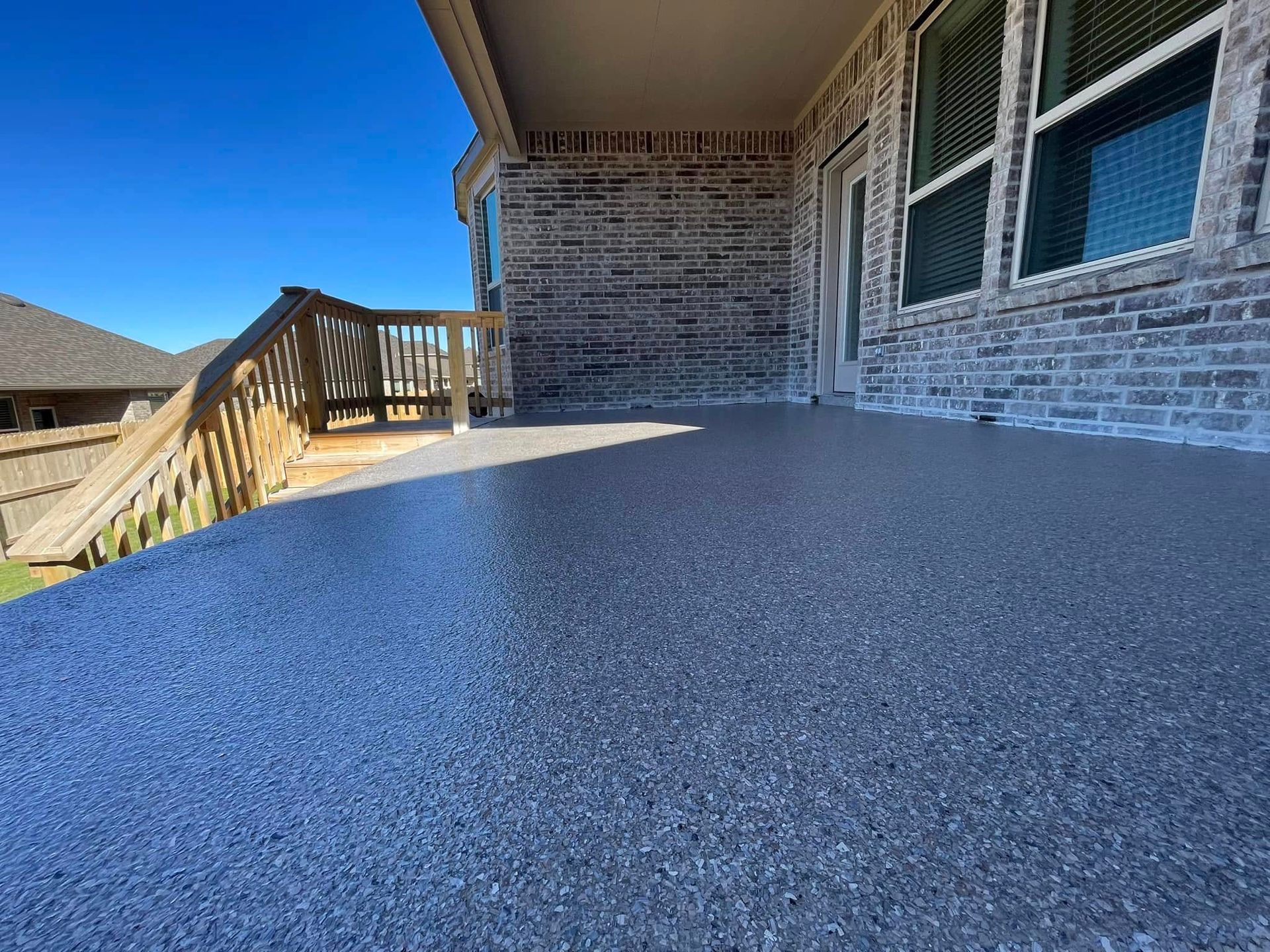 Gray, speckled concrete patio with wooden railing, brick facade, and clear blue sky.
