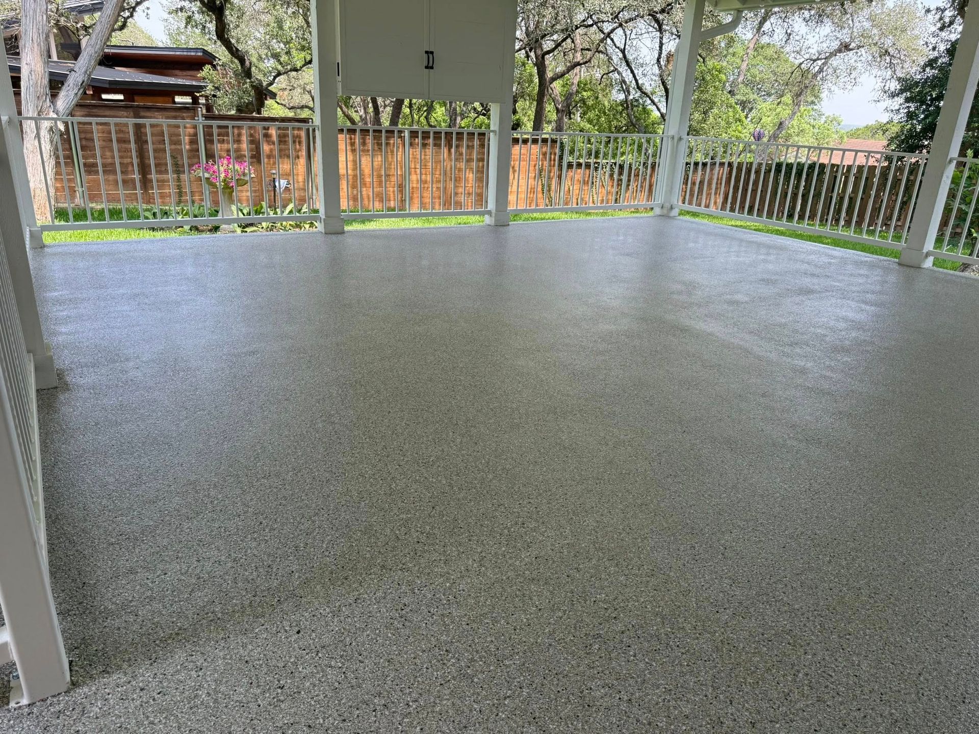 Covered outdoor patio with speckled gray floor; white railing and supports, wooden fence in the background.
