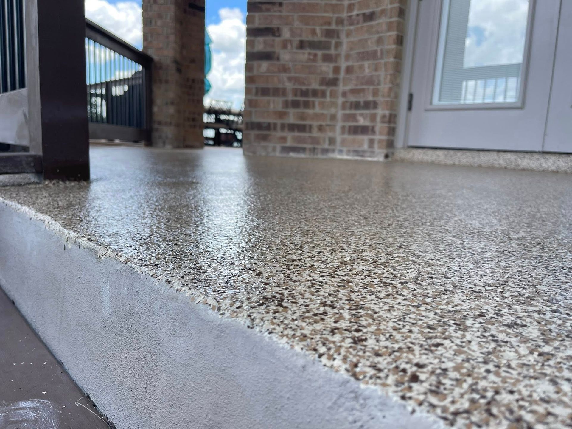 Close-up of a newly-coated concrete patio with a speckled pattern, near a brick wall, a door, and a railing.