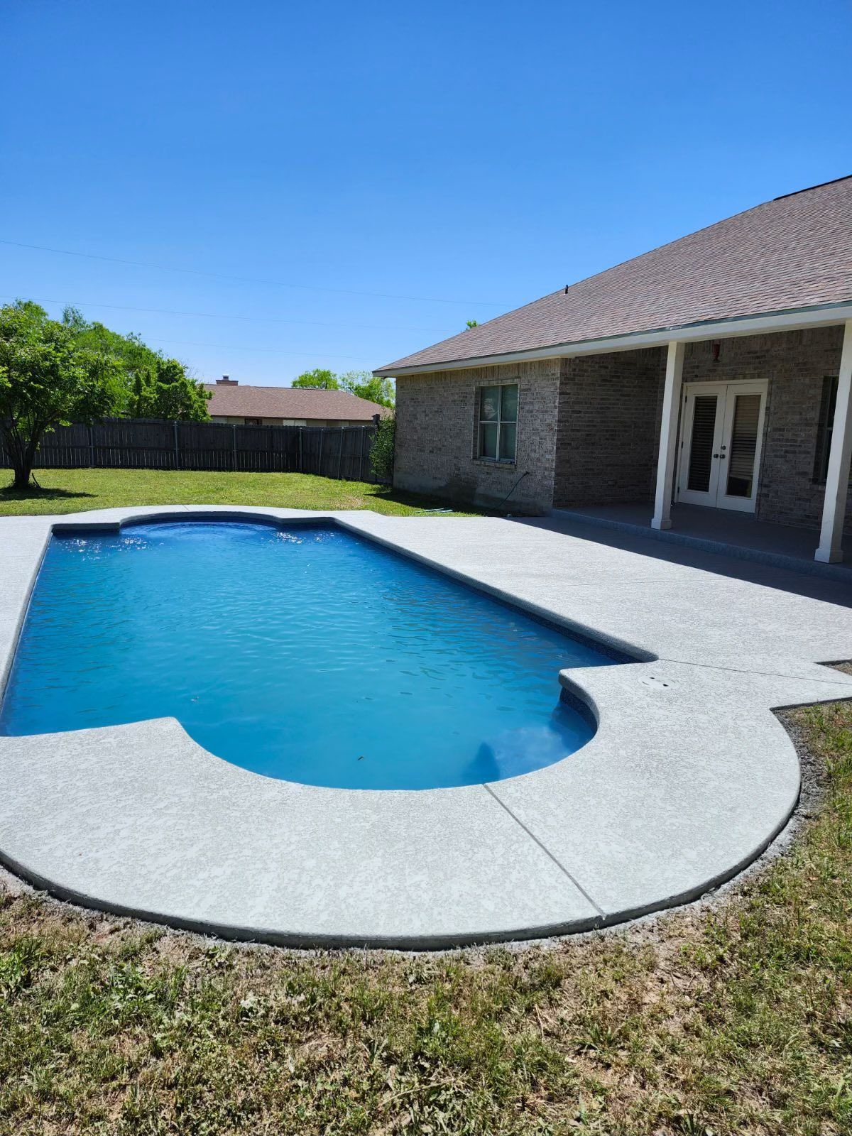 Pool with blue water and gray concrete patio next to a brick house and grassy yard.
