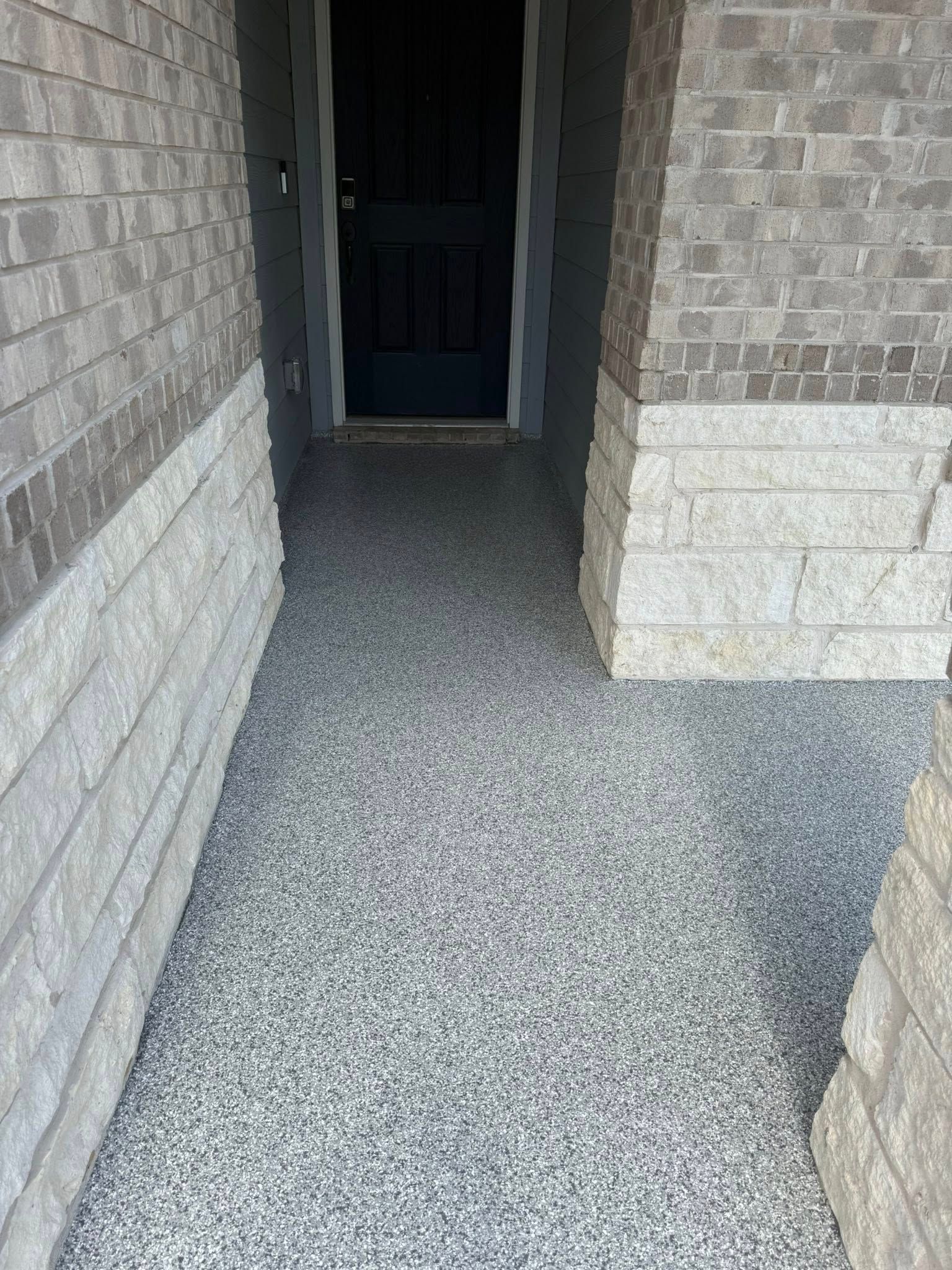 Exterior walkway with speckled gray flooring, brick and stone walls, and a dark front door.