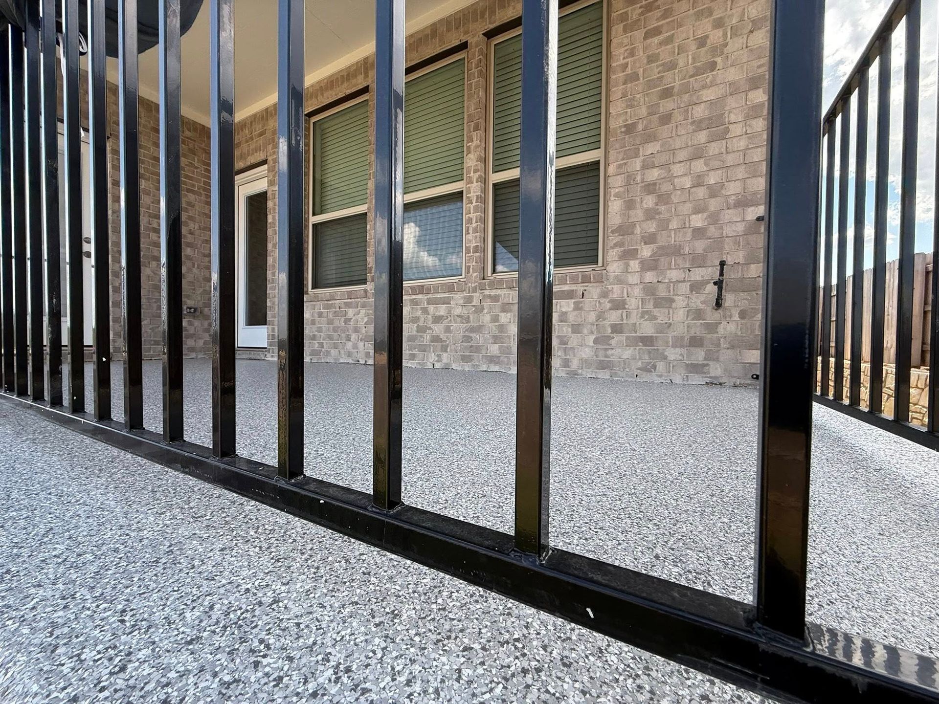 Black metal fence on a gray speckled patio, leading to a brick-walled house with windows.