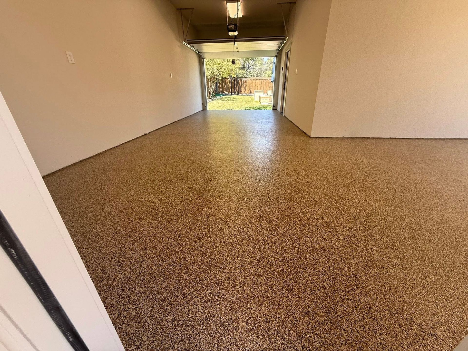 Garage interior with speckled brown floor and open door to a backyard.