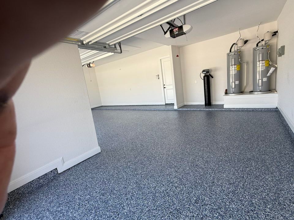 An empty garage with a speckled gray floor, water heaters, and a black tank against the white walls.
