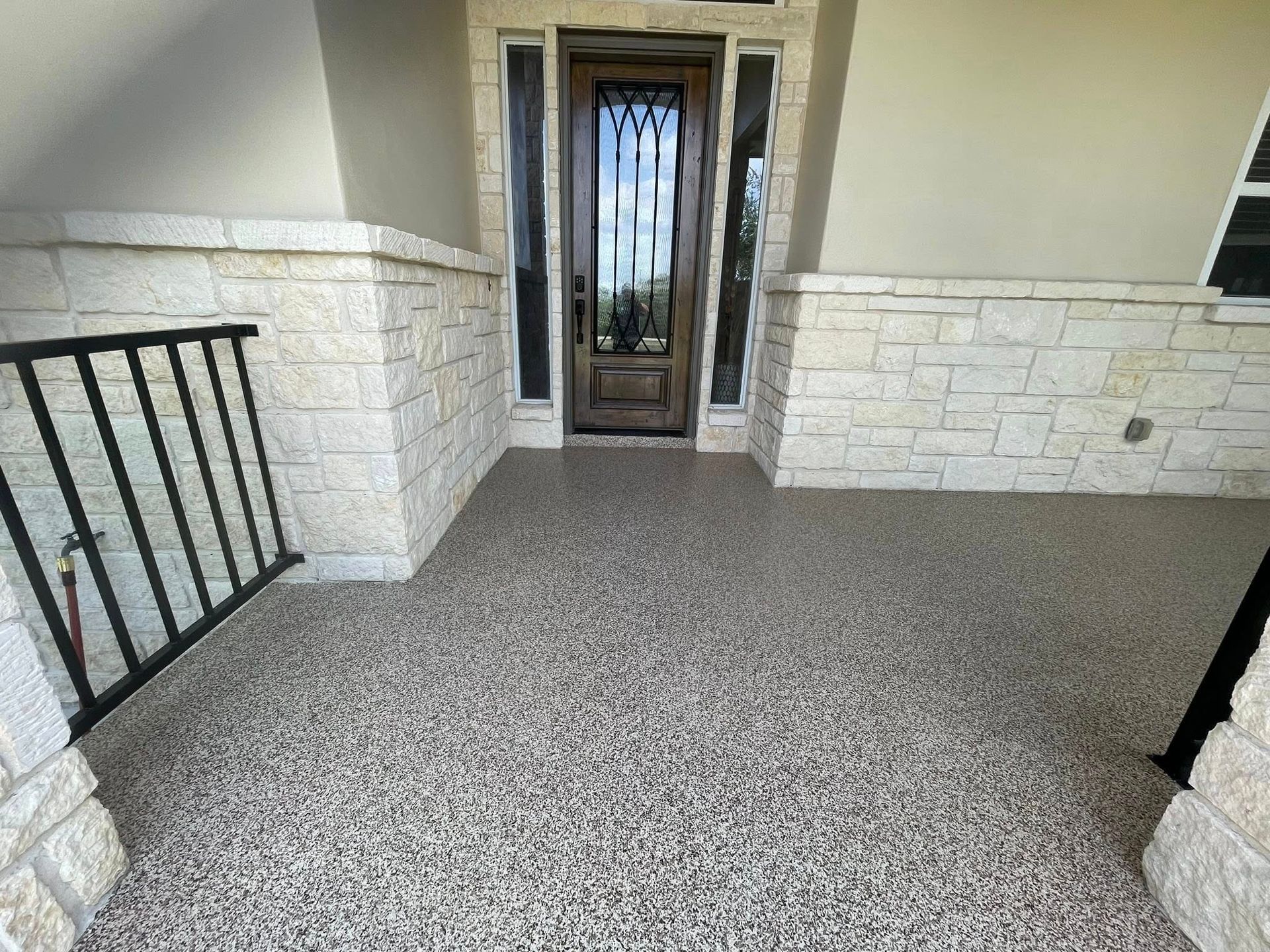 Covered porch with speckled gray flooring, light stone walls, and a dark wood door.