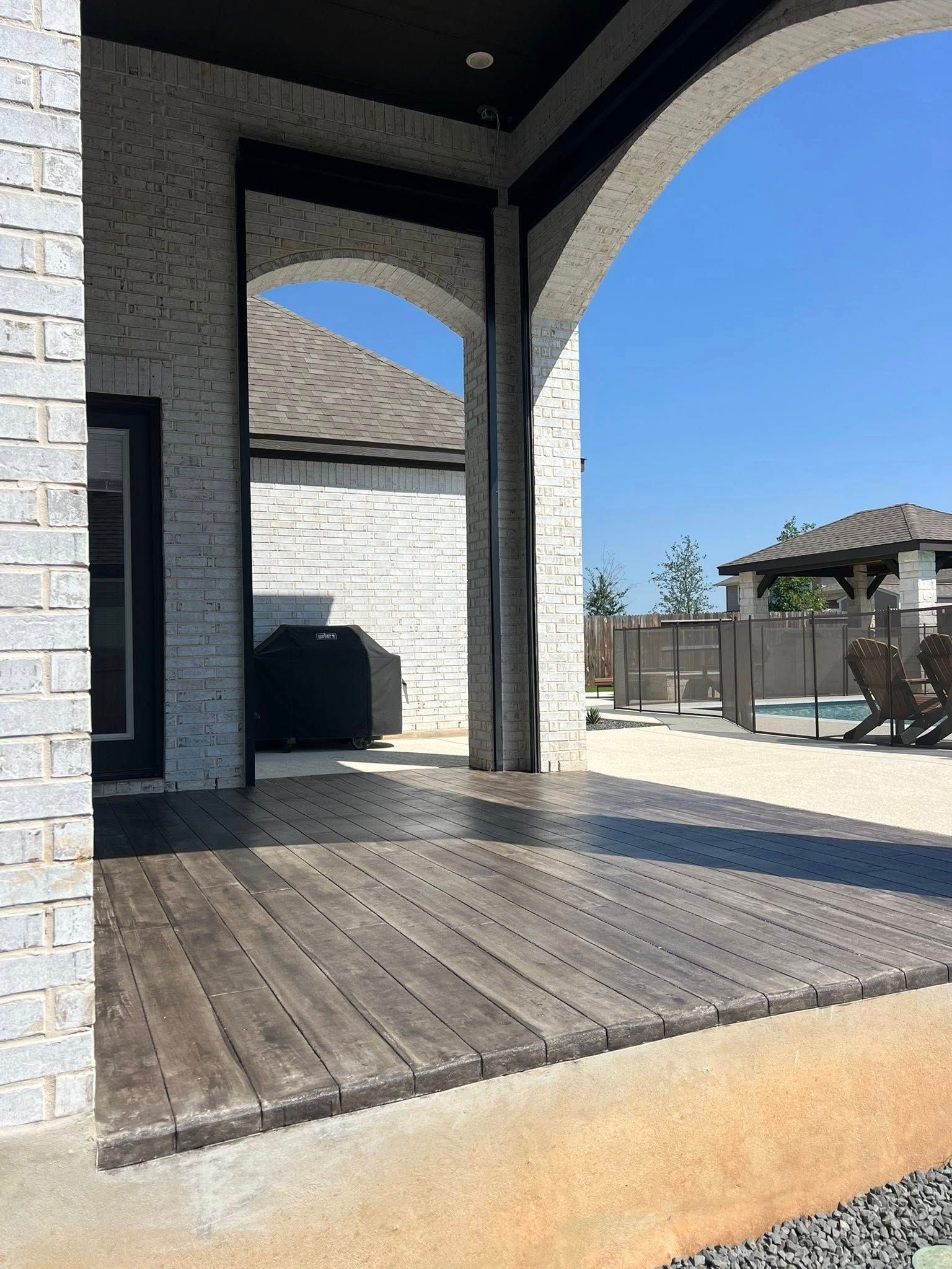 Outdoor patio with a wood-like floor, brick walls, and a view of a pool area on a sunny day.