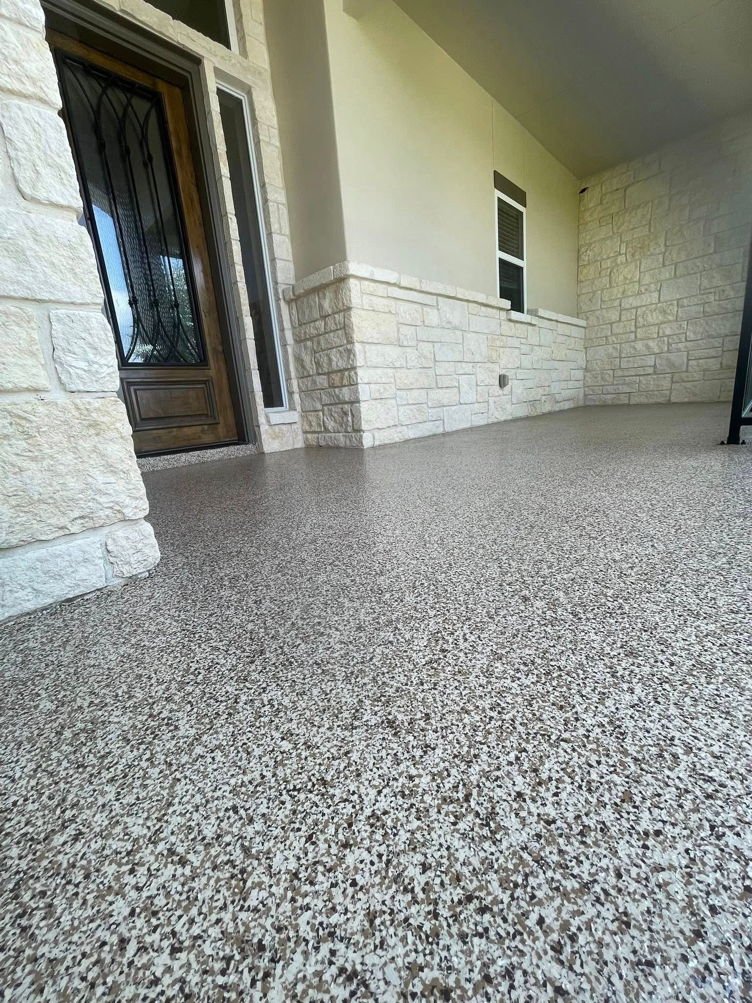 Low-angle view of a covered porch with a speckled, beige and brown floor. Stone pillars and walls frame the entrance.