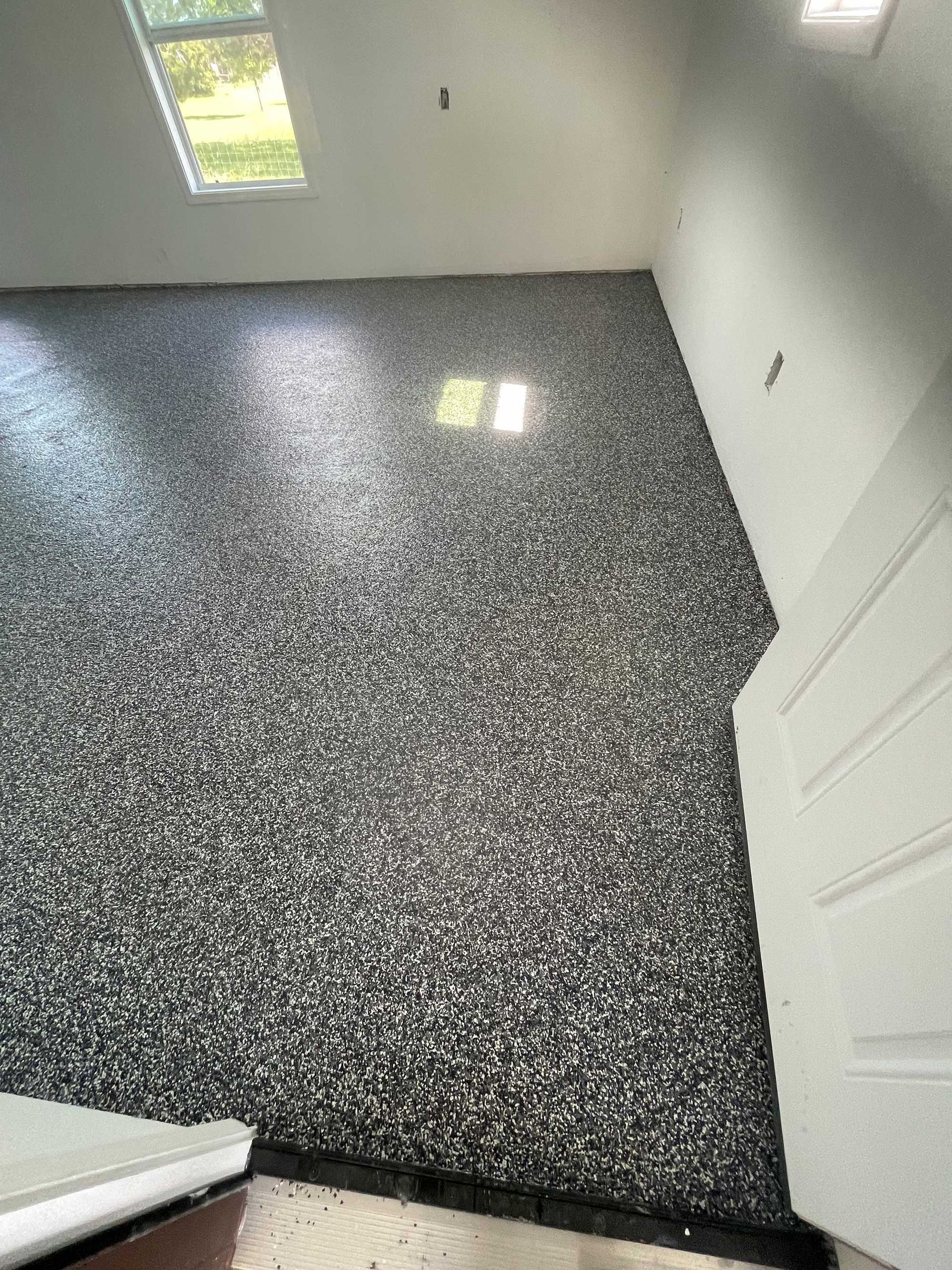 Black, gray, and white speckled epoxy flooring in a room with a window and white walls.