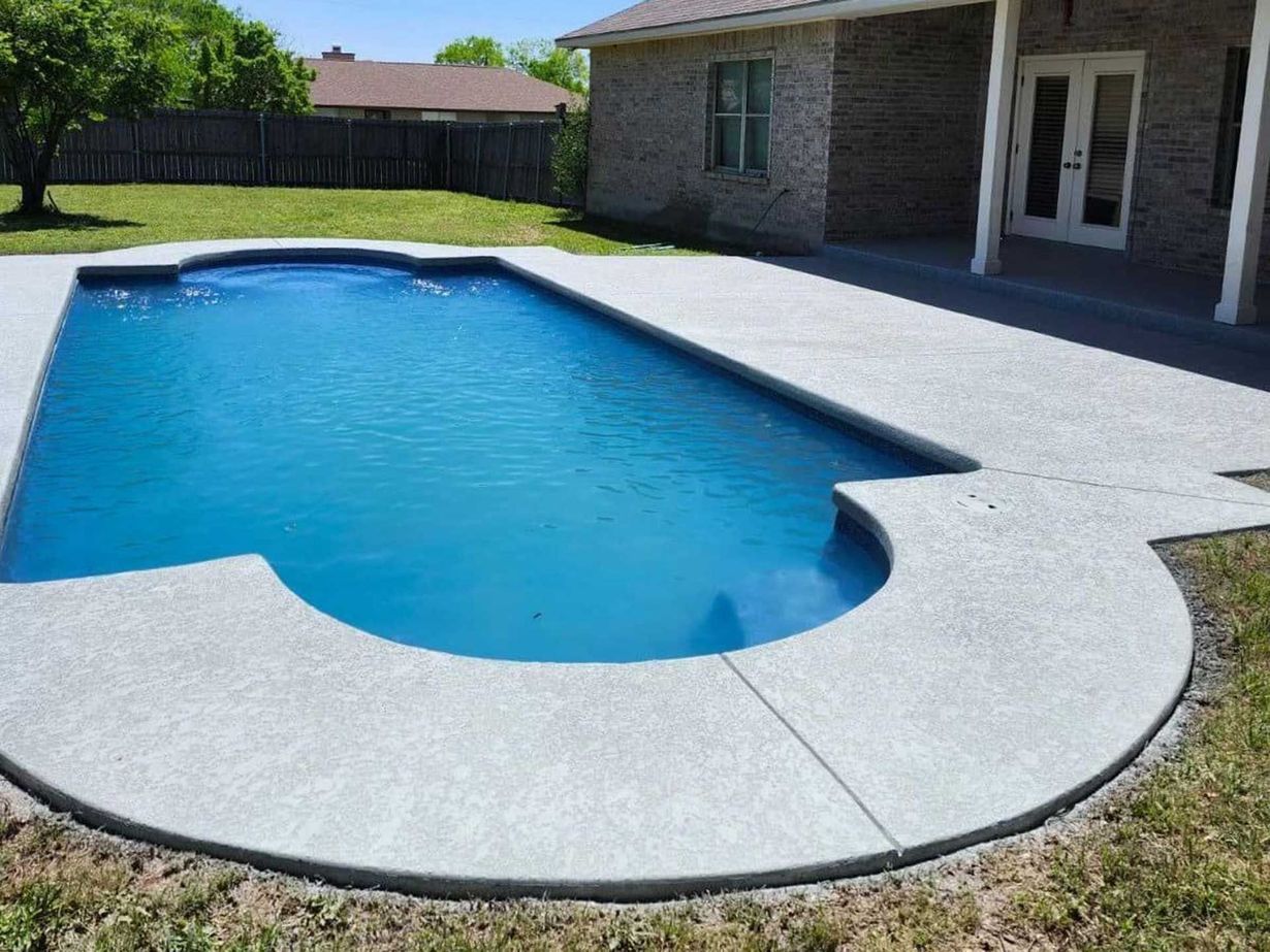 A blue-water swimming pool with a concrete patio, located in a backyard next to a brick house.