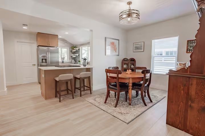 Bright dining room with a round wooden table, chairs, light wood flooring, and an open kitchen beyond.