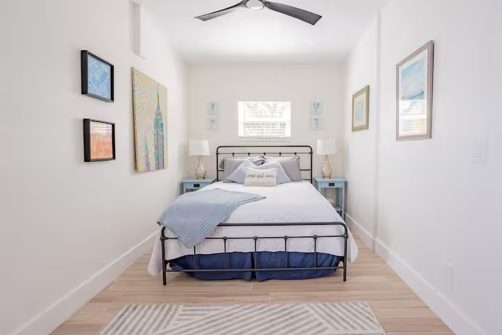 Bright bedroom with a metal bed, blue bedding, wall art, and a ceiling fan near a window.
