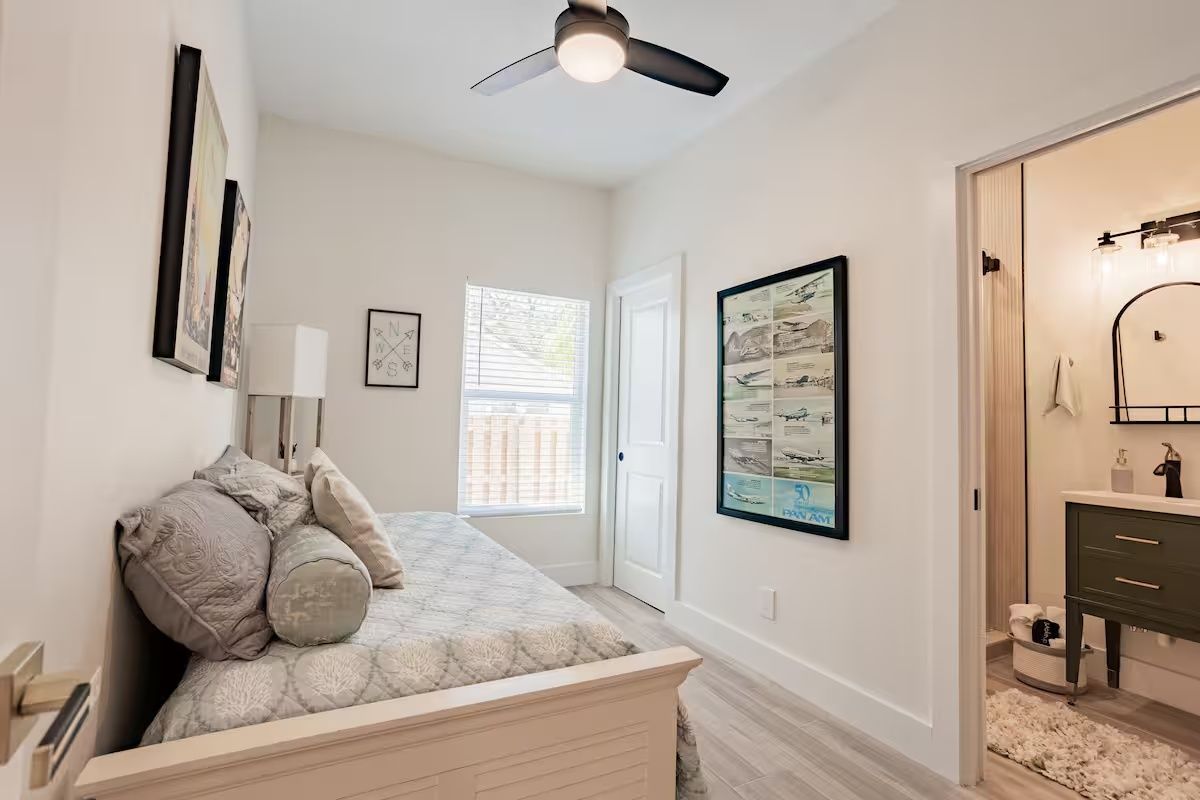 Bright bedroom with a bed, ceiling fan, framed art, and an open doorway to a bathroom.