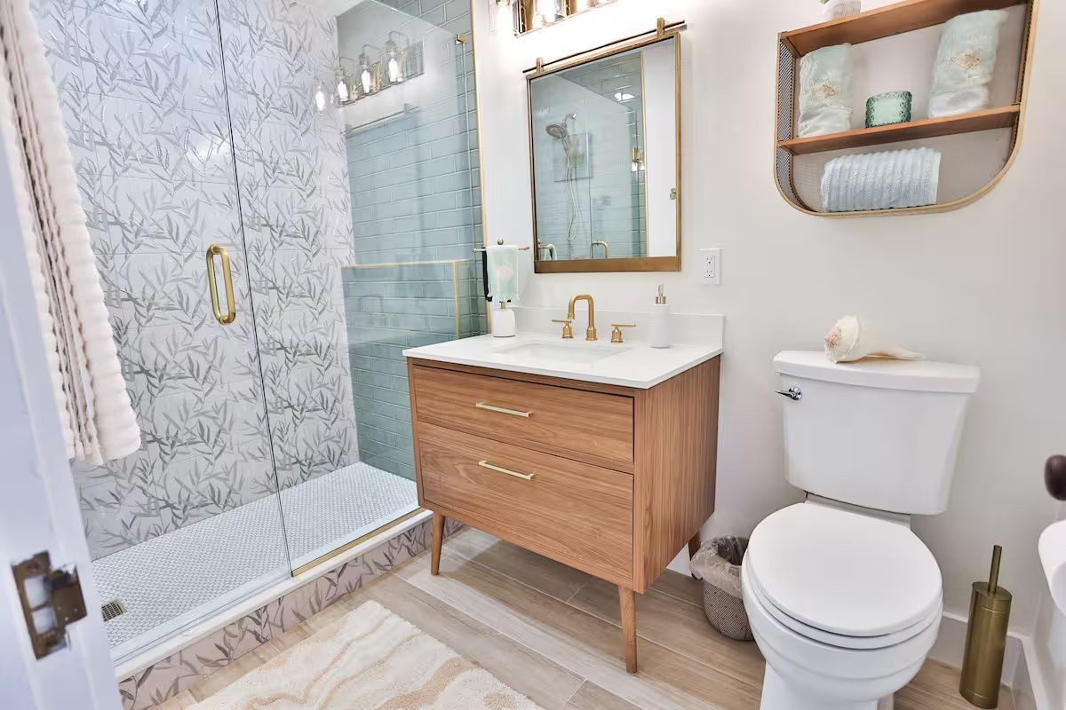 Bright modern bathroom with glass shower, wood vanity, white toilet, and patterned tile floor