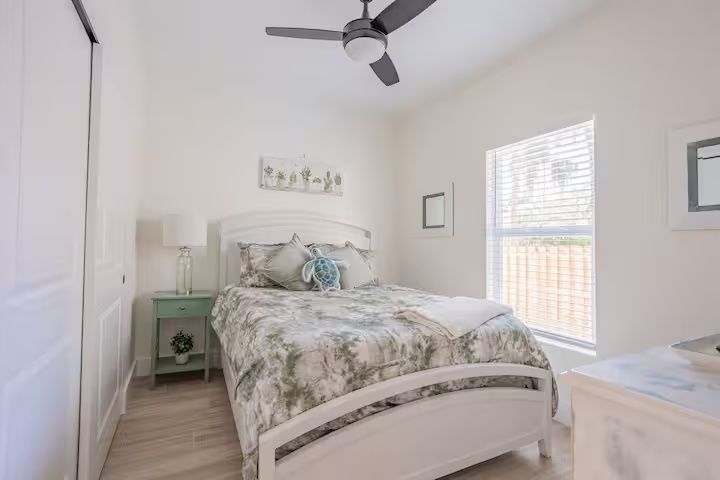 Bright bedroom with white walls, a bed, ceiling fan, and window blinds in soft natural light