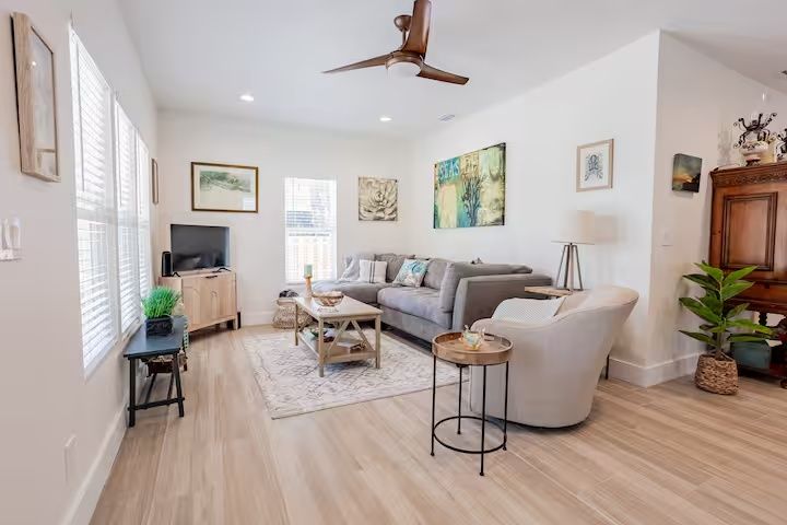 Bright living room with gray sofa, white armchair, coffee table, plants, and ceiling fan.