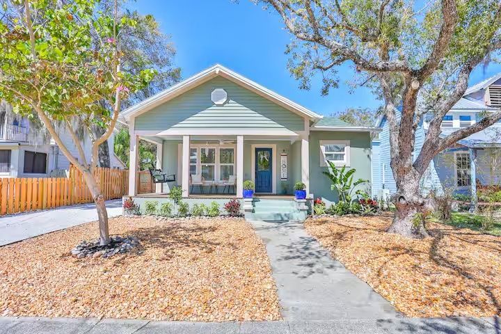 Small green bungalow with a front porch, blue door, and a walkway lined by dry landscaping.