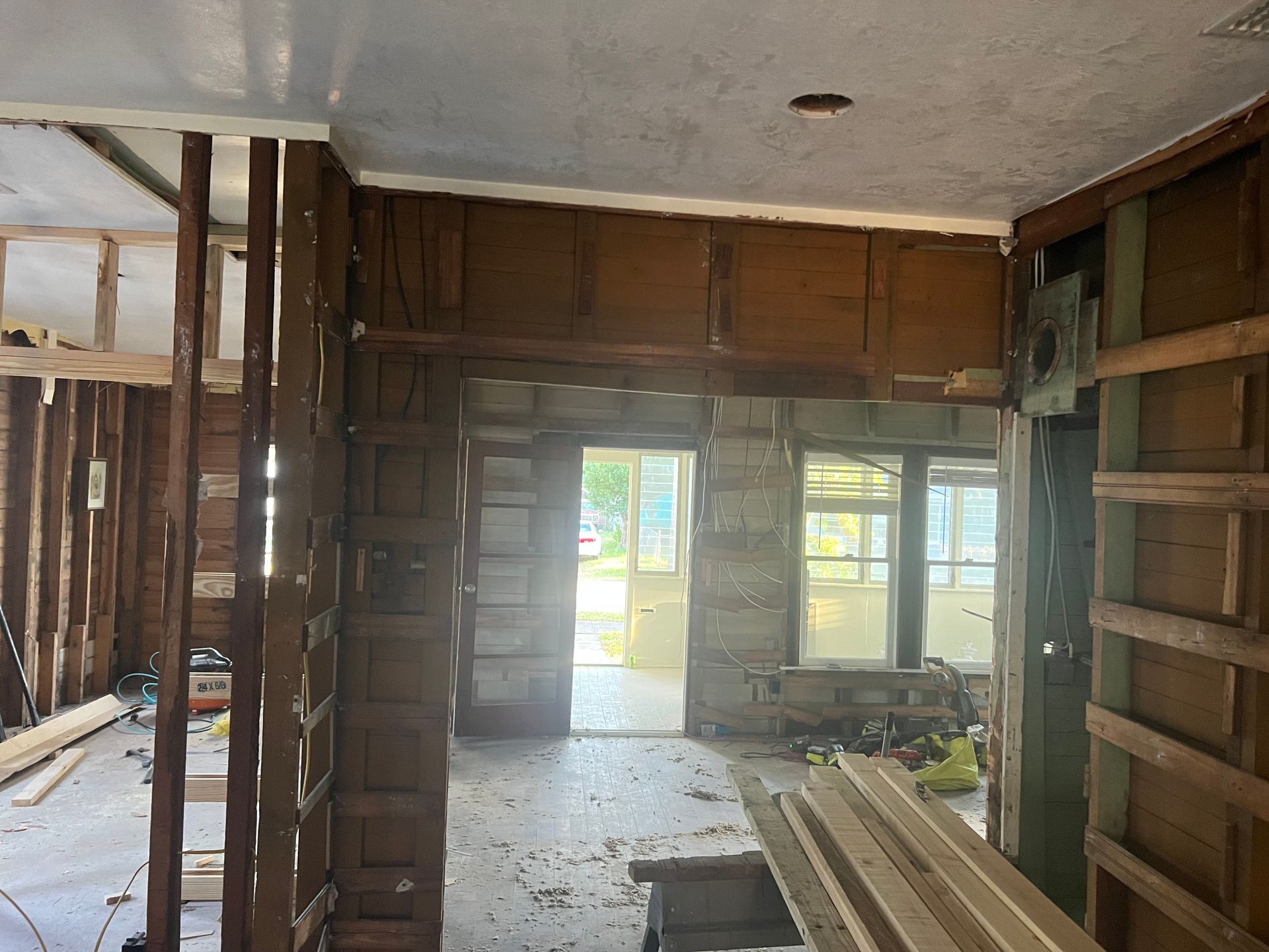 Damaged interior room under renovation with exposed studs, debris on the floor, and bright windows and doorway.