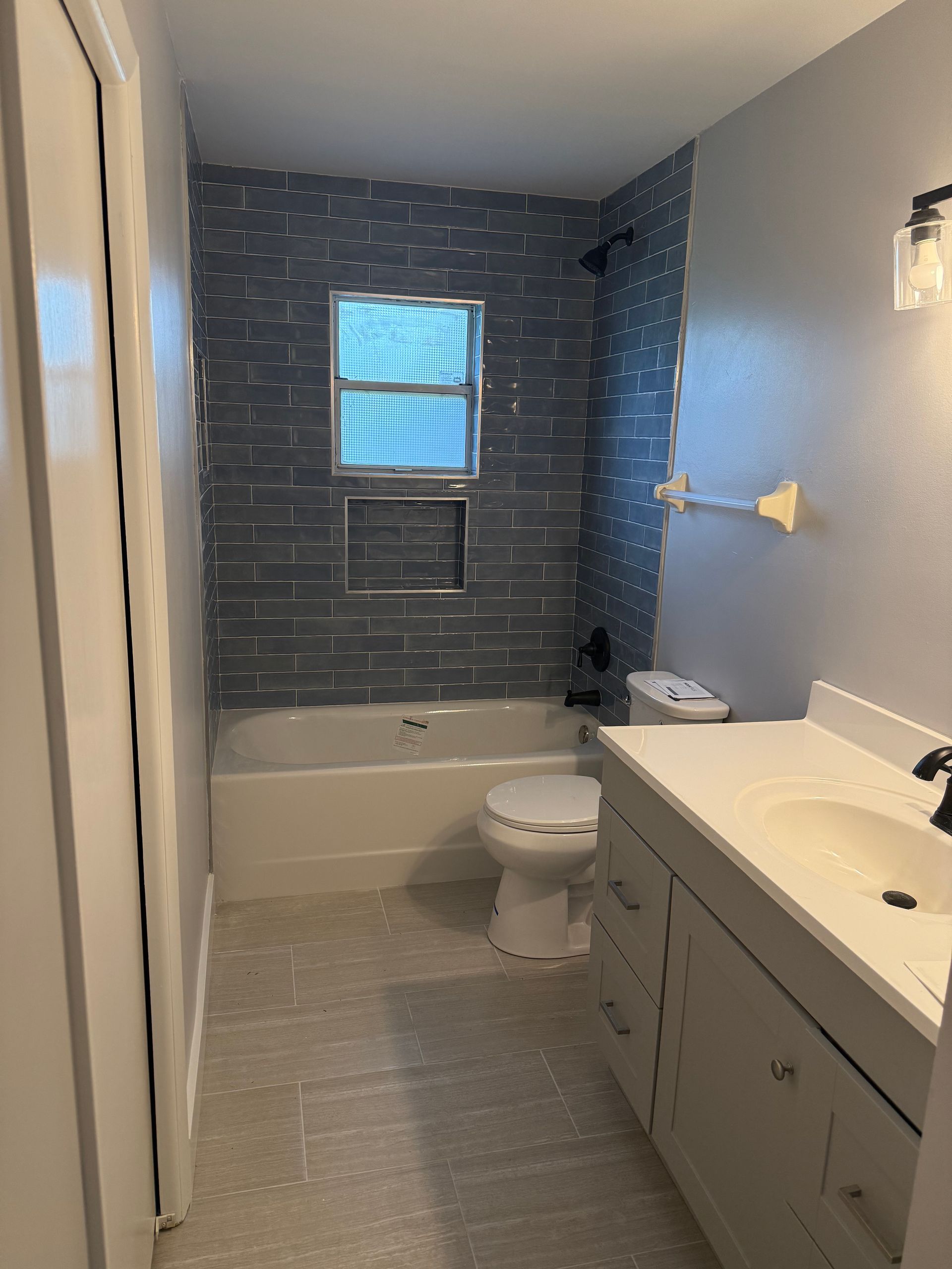 Compact bathroom with gray tile, white tub and toilet, blue accent wall, and vanity sink.