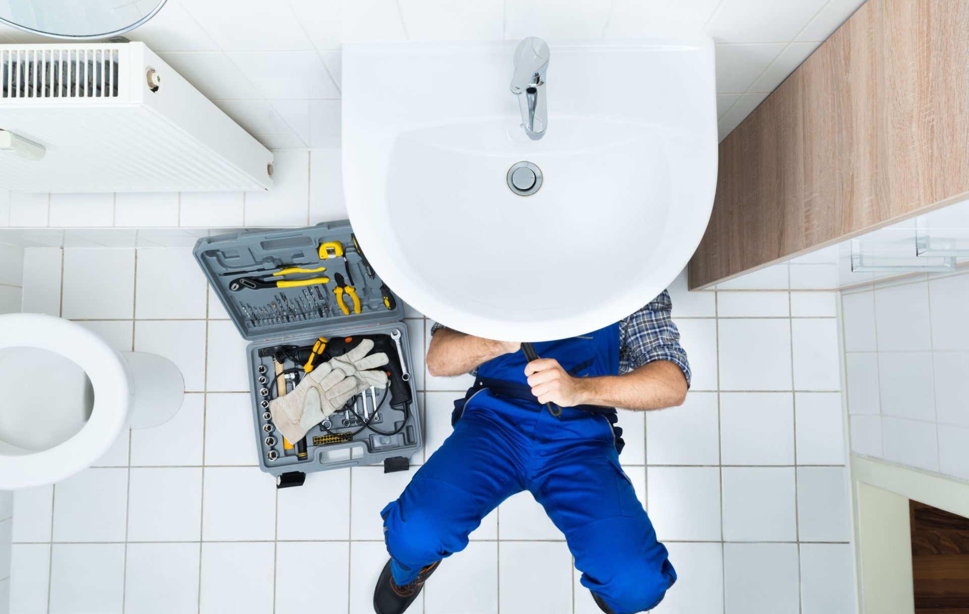 Plumber in blue overalls working under a white sink with tools, gray toolbox, and toilet in a white tiled bathroom.