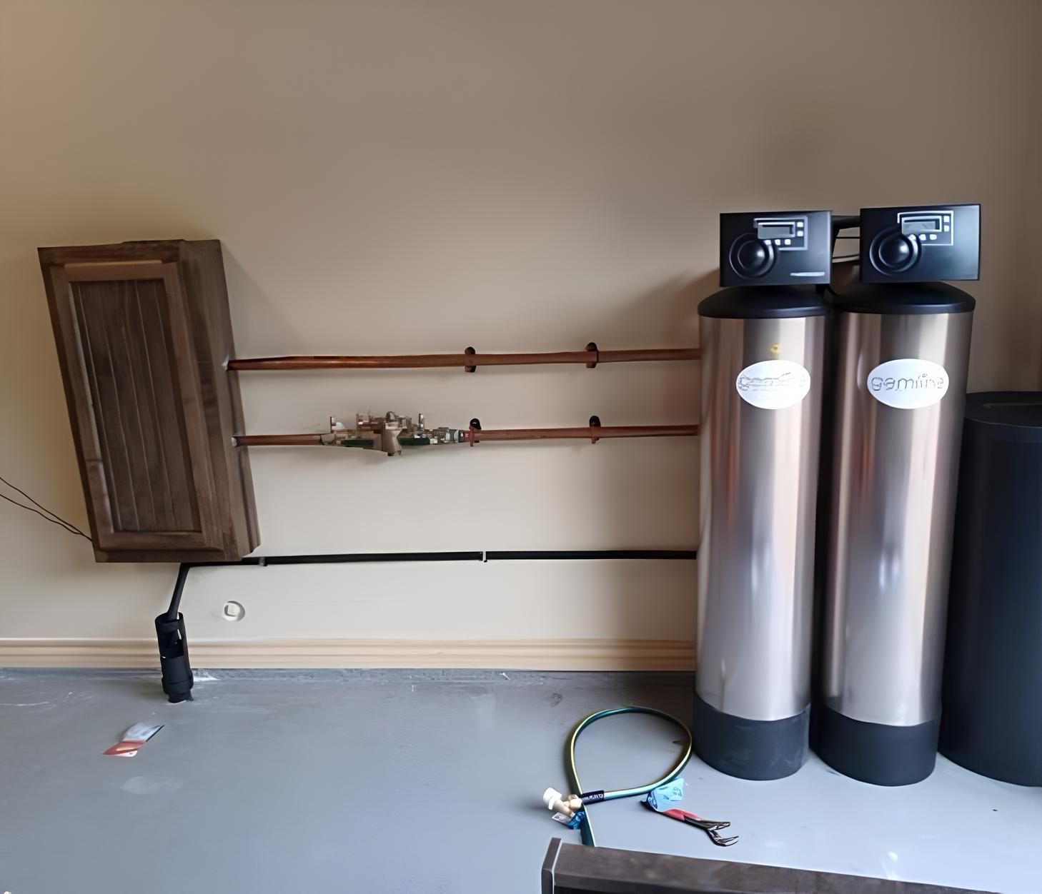 Water filtration system with two stainless steel tanks, copper pipes, and a wooden cabinet on a gray floor against a beige wall.