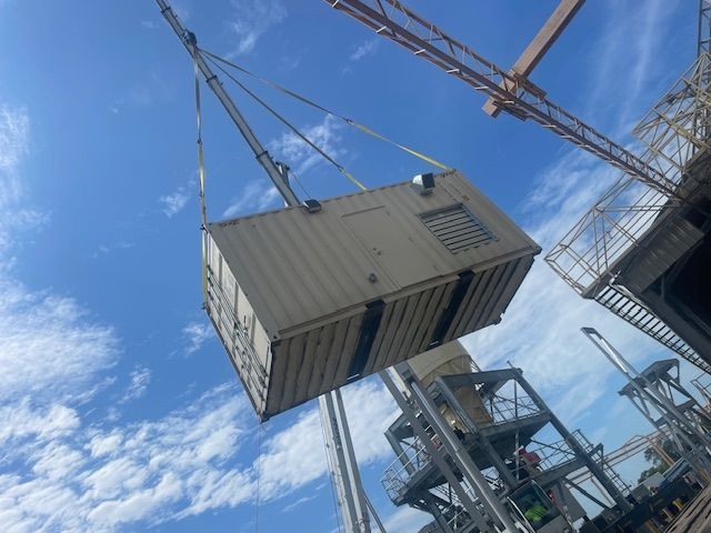 A large beige container is lifted by a crane on a construction site under a blue sky.