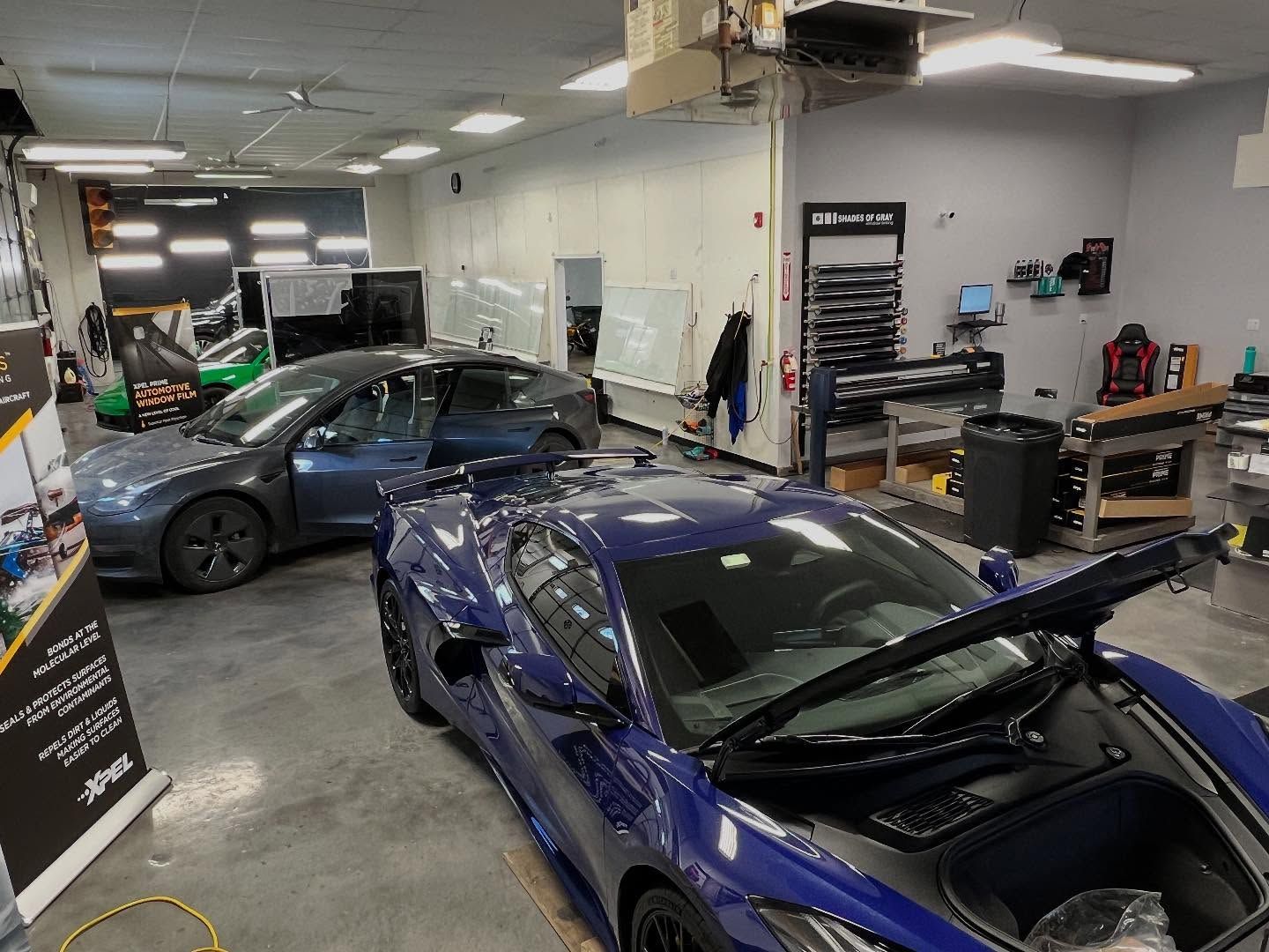 Inside a car detailing shop: Blue sports car with hood open, Tesla, and green car are parked.