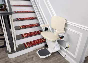 Beige stairlift beside carpeted stairs with red runner and wall-mounted controls