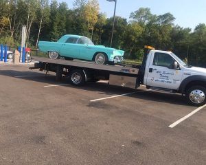 A teal vintage car is loaded onto the flatbed of a white All Seasons Towing truck in an outdoor parking lot.