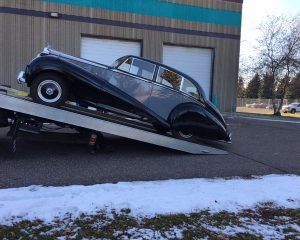 A vintage black sedan car being loaded onto a flatbed tow truck in front of a building on a cold day.