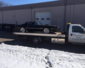 A black muscle car loaded onto the flatbed of a tow truck in front of a building with snow on the ground.