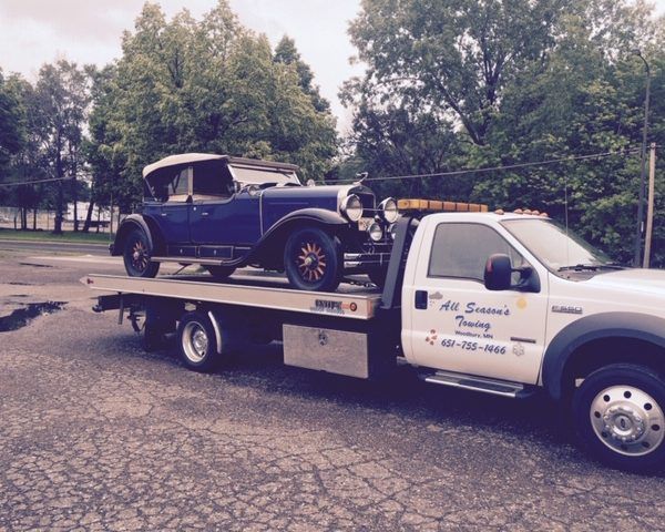 A vintage blue convertible car loaded onto the flatbed of an All Season's Towing truck outdoors.