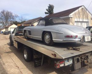 A silver Mercedes-Benz 300SL Roadster sits on the flatbed of a tow truck parked on a residential street.
