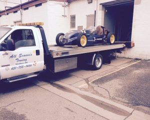A white flatbed tow truck from All Season's Towing transporting a vintage dark blue race car to a building loading dock.