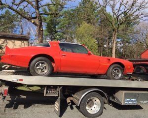A bright red classic Camaro muscle car secured on the bed of a flatbed tow truck outdoors on a sunny day.