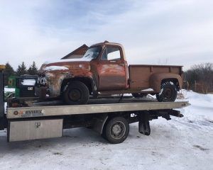 A rusted vintage pickup truck sits on the back of a flatbed tow truck in a snowy landscape.