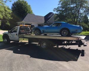 A bright blue classic Chevrolet Corvette sports car loaded onto a white flatbed tow truck parked on a residential street.