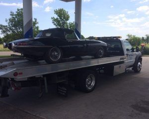 A black vintage Jaguar E-Type convertible is secured on the flatbed of a tow truck at a gas station.