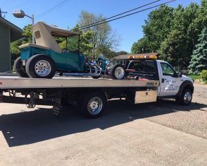 A turquoise classic hot rod car is being transported on the flatbed of a white tow truck on a sunny, paved lot.