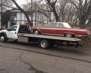 A white flatbed tow truck transporting a vintage, two-tone red and white classic car along a street.