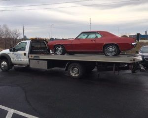 A red vintage Camaro sits on the bed of a white flatbed tow truck in a parking lot.