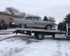 A silver classic Chevrolet car is being transported on the flatbed of a tow truck in a snowy, outdoor parking lot.