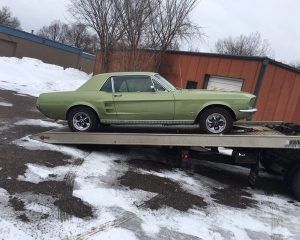 A light green vintage Ford Mustang coupe secured on a flatbed tow truck in a snowy, outdoor lot.