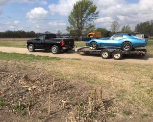 A black pickup truck tows a trailer carrying a bright blue vintage sports car on a dirt path in a grassy field.