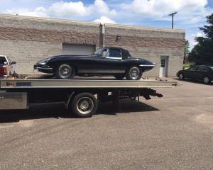 A black Jaguar E-Type convertible is secured on the bed of a flatbed tow truck in a paved lot.