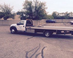 A white flatbed tow truck parked in an empty asphalt lot with trees and a fenced background.