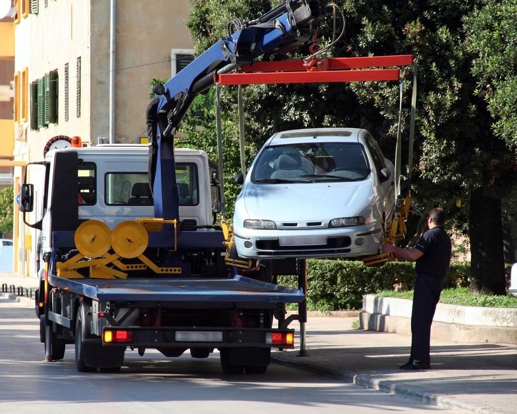 A tow truck crane lifts a silver car off the street as a uniformed officer stands nearby.