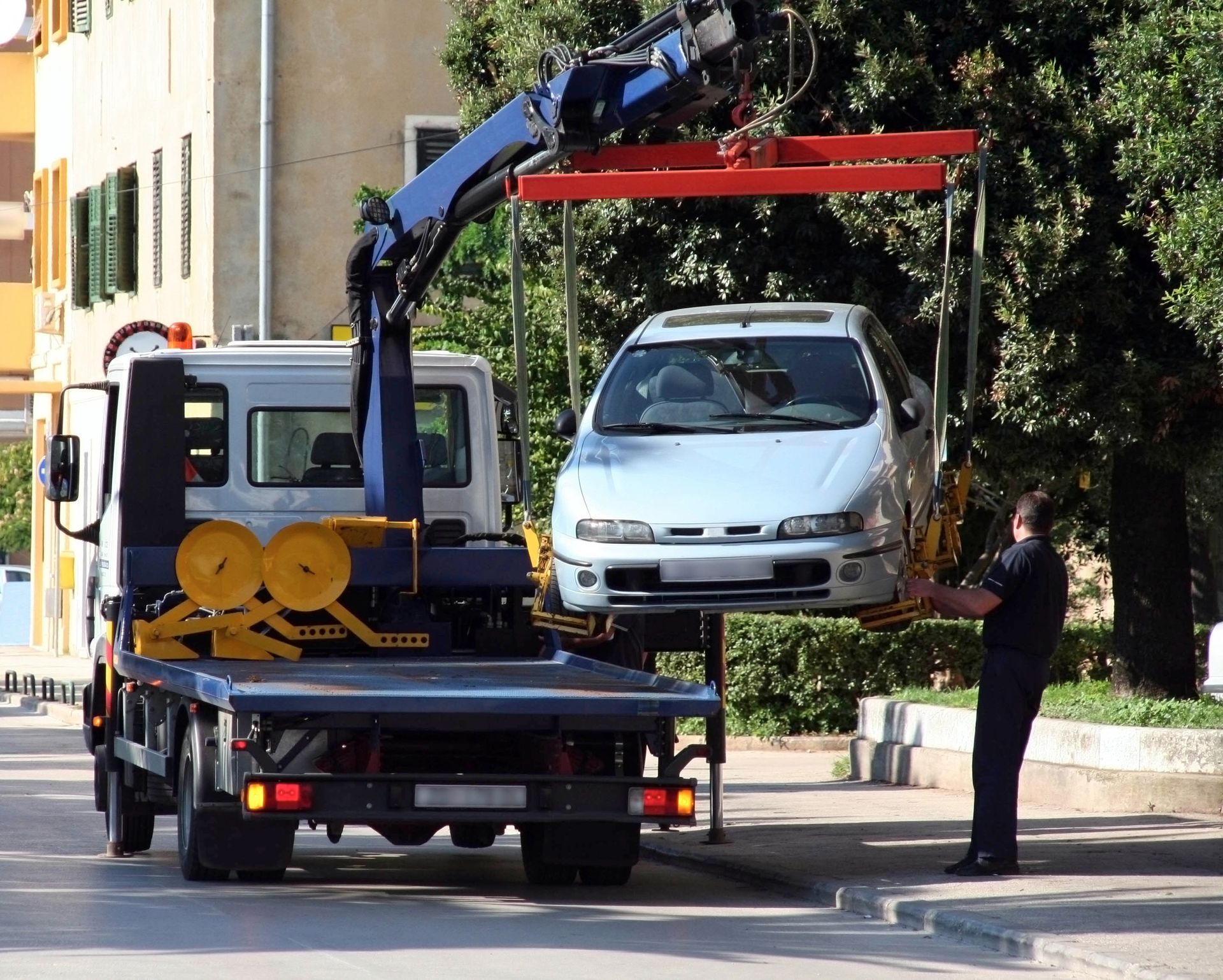 A tow truck crane lifts a silver car off the street as a uniformed officer stands nearby.